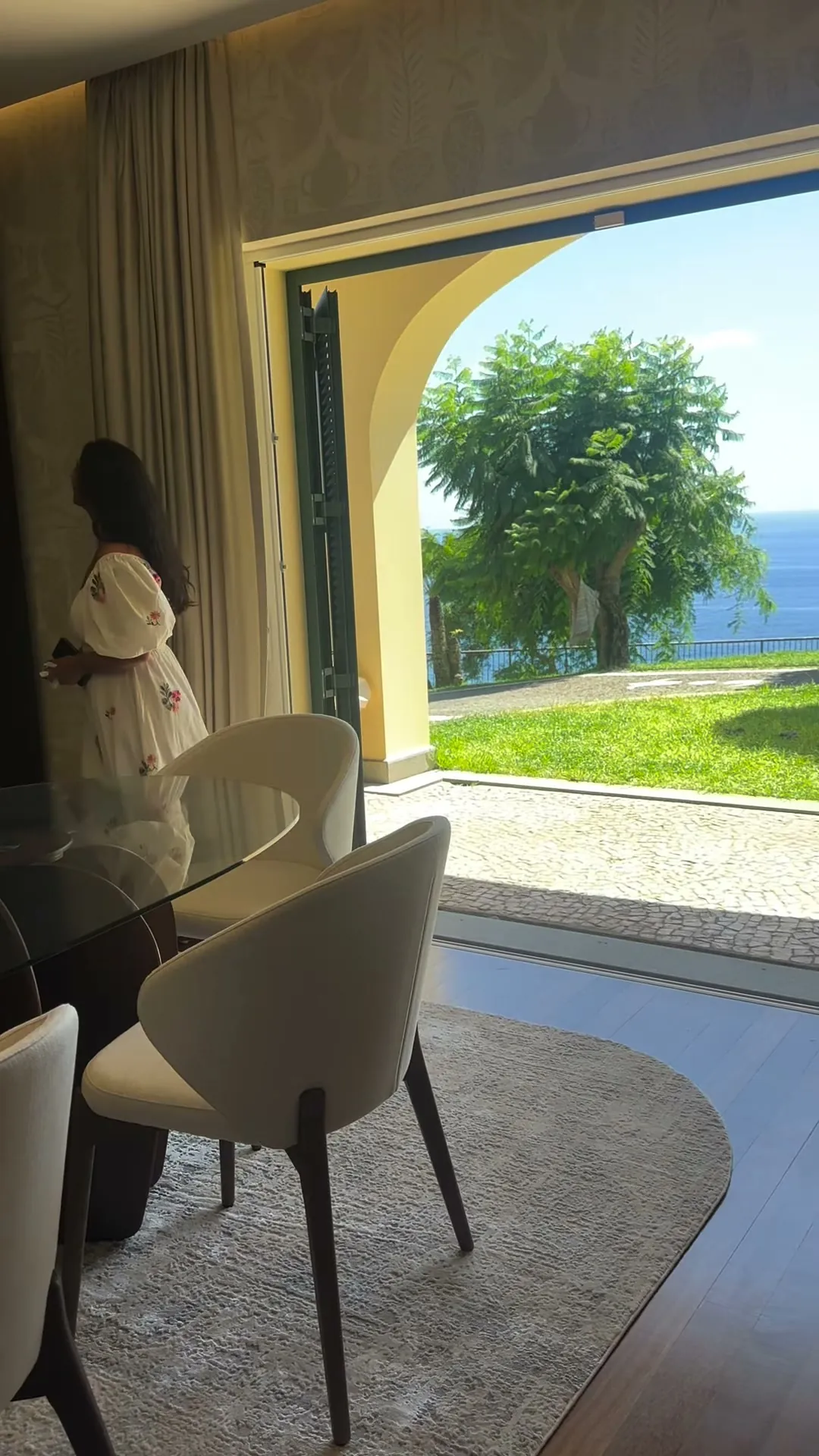 Sala de jantar de uma moradia com arco aberto para o jardim e o mar, cadeiras claras e mesa de vidro